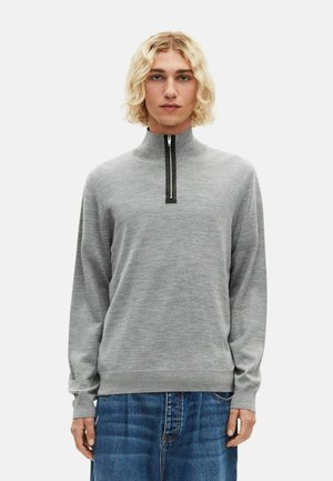 Giovane con capelli biondi fino alle spalle, che indossa un sweater grigio con mezza zip e jeans blu, in piedi davanti a uno sfondo neutro.