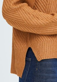 Pull en maille côtelée texturée de couleur marron chaud, avec des fentes sur les côtés et une coupe décontractée, associé à un jean en denim bleu foncé.