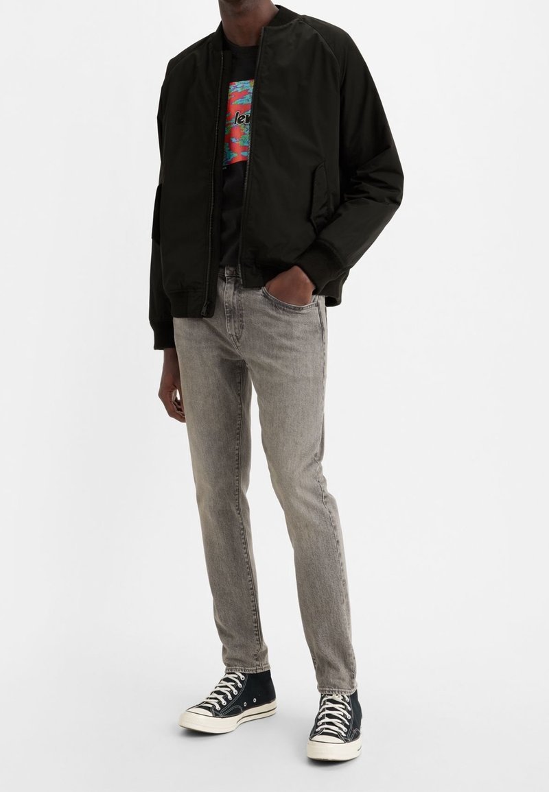 Black bomber jacket, graphic t-shirt, light grey jeans, and black high-top sneakers. The jacket features a zip closure and ribbed cuffs.