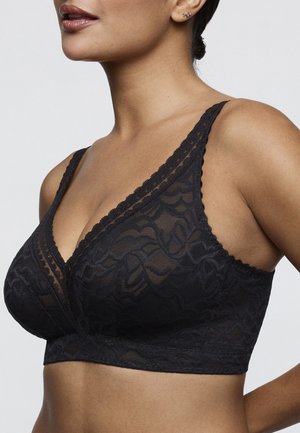 Woman wearing a black lace bralette with thin straps and floral patterns, shown from shoulders to midriff against a plain background.