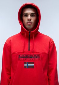Felpa rossa con cappuccio in tessuto morbido, con zip a quarto di lunghezza, logo nero e un patch con una bandiera. Il cappuccio è foderato e dotato di coulisse regolabili.