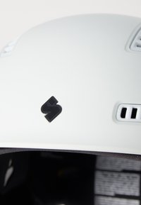 Biały kask z otworami wentylacyjnymi, czarnym logo z przodu i miękką wewnętrzną wyściółką. Charakteryzuje się gładką, matową fakturą i delikatnym wygięciem.
