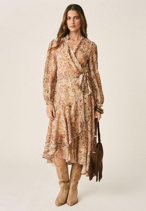 Femme portant une robe portefeuille à manches longues avec motif cachemire et volants, des bottes hautes en daim beige, et tenant un sac à main marron à franges.