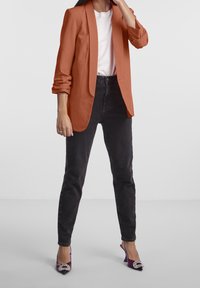 Rostfärgad blazer över en vit t-shirt, parat med mörka jeans och lila klackar med en juvelaccent. Slät tygkvalitet.