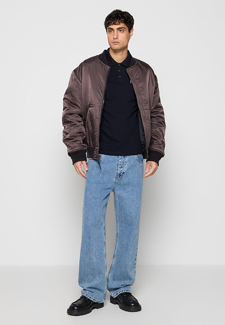 Hombre con chaqueta bomber de satén marrón, polo negro, vaqueros de pierna ancha azul claro y zapatos negros, de pie sobre fondo blanco.