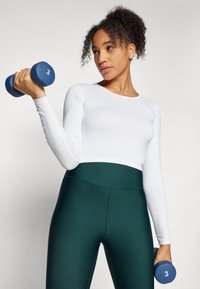 Long-sleeve white top paired with high-waisted dark green leggings. Model holds blue dumbbell with the number 3 printed on it.