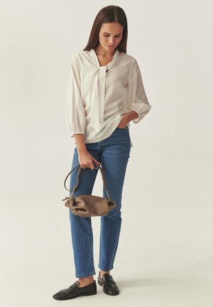 Naine valges pluusis ja sinistes teksades, hoides ühes käes pruuni käekotti ja teist kätt taskus, seistes ühevärvilise tausta ees.