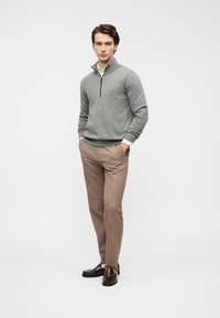 Ανοιχτό γκρι πουλόβερ με φερμουάρ στο λαιμό και ριμπ μανσέτες, σε συνδυασμό με ανοιχτό καφέ παντελόνι. Το μοντέλο στέκεται με τα χέρια στις τσέπες, φορώντας σκούρα καφέ μοκασίνια.