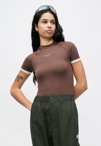 Καφέ πουκάμισο με κοντά μανίκια και ψηλό γιακά, με άσπρες μανσέτες και μικρό λευκό λογότυπο Nike στο στήθος. Φορεμένο με ελαιδί παντελόνια.