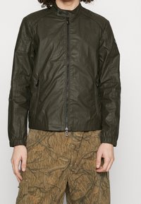 Olivgrön, lättviktsjacka med ståkrage, främre dragkedja och två sidofickor; har elastiska ärmslut och detaljerad sömnad.