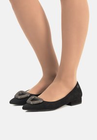 Bailarinas de ante negro con punta afilada, que presentan una hebilla metálica decorativa en la parte superior y un tacón bajo tipo bloque.