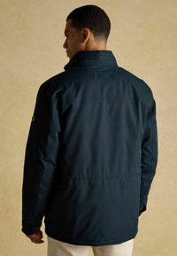 Muž v tmavomodrom saku s vysokým golierom, zobrazený zozadu, stojací proti jednému svetlému béžovému pozadiu.