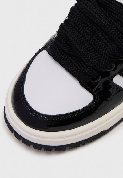 Black patent leather sneaker with white toe cap and thick black laces. Features textured tread and smooth white accents.