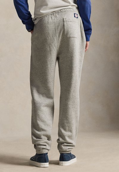 Person som har på seg lysegrå joggebukser med elastiske mansjetter og en baklomme, kombinert med marineblå joggesko og en blå langermet skjorte.