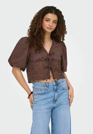 Młoda kobieta o kręconych włosach, ubrana w brązowy krótki top z koronką i bufkami oraz jasnoniebieskie, wysokiej talii dżinsy, pozująca z ręką na biodrze.