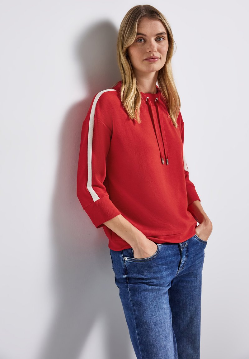 Pullover rosso con cappuccio, caratterizzato da strisce laterali bianche, maniche a 3/4 aderenti e una vestibilità morbida, abbinato a jeans in denim blu.