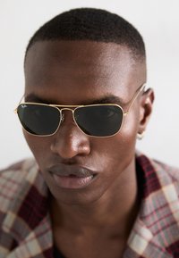 Goldfarbene Sonnenbrille mit dunklen Gläsern. Der Stil zeichnet sich durch ein schlankes Design aus und wird von einem Modell vor einem neutralen Hintergrund getragen.