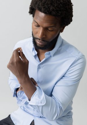 Camisa de manga larga de color azul claro, hecha de una tela suave con puños de botón, combinada con jeans de mezclilla oscura. El cuello es clásico y estructurado.
