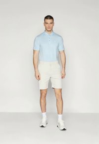 Camisa polo azul claro y pantalones cortos color blanco sucio con bolsillos. El modelo lleva zapatillas blancas y calcetines cortos, de pie sobre un fondo liso.