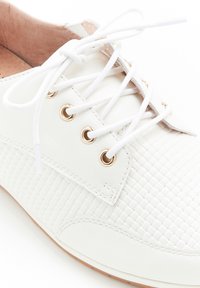 Sneaker bianco realizzato in materiale sintetico testurizzato, con motivo intrecciato, occhielli dorati e lacci bianchi. La soletta marrone è visibile.