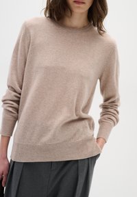Pull en cachemire beige avec un col rond et des poignets côtelés. Texture douce avec une coupe décontractée. Porté sur un pantalon gris plissé.