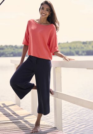 Lächelnde Frau in korallenrotem Oberteil und marineblauer, weiter Hose barfuß auf einem hölzernen Steg am ruhigen Wasser, die sich an ein weißes Geländer lehnt, im Hintergrund grüne Bäume.