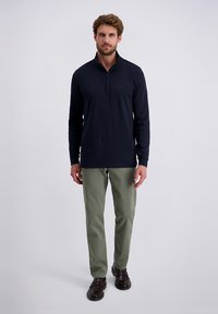 Marineblauwe longsleeve polo met halfrits, gecombineerd met lichtgroene broeken en donkerbruine schoenen. Model staat tegen een effen achtergrond.