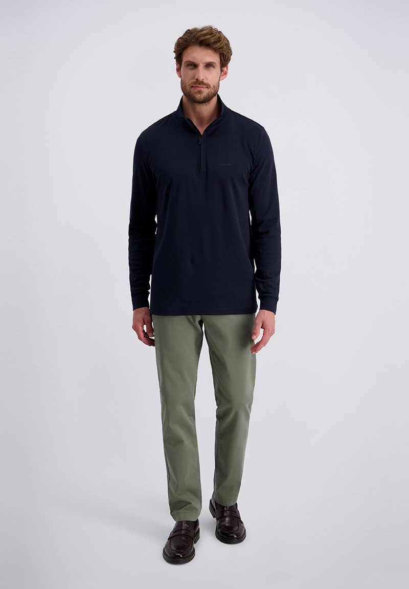 Marineblauwe longsleeve polo met halfrits, gecombineerd met lichtgroene broeken en donkerbruine schoenen. Model staat tegen een effen achtergrond.