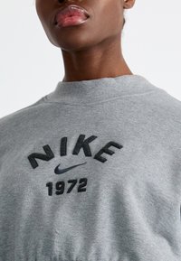 Pelēks džemperis ar apaļu kakla izgriezumu, ar reliefu izteiksmīgu "NIKE" logo un "1972" tekstu melnā krāsā. Mīksta auduma izstrādājums ar elastīgu apakšmalu.