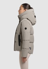Veste rembourrée gris clair avec une grande capuche, dotée d'une poche zippée et d'un patch logo noir sur la manche. Texture lisse et matelassée.