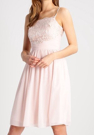 Frau trägt ein hellrosa ärmelloses Kleid mit Spitzenoberteil und einem fließenden Rock, die Hände sanft vor sich gefaltet.