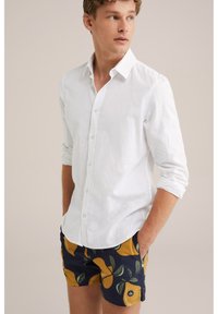 Weißes Leinenhemd mit Knöpfen und hochgerollten Ärmeln, kombiniert mit marineblauen Badeshorts, die ein Muster aus gelben Birnen und grünen Blättern zeigen.