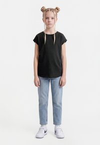 Noor tüdruk seistes, seljas must t-särk, helesinised teksad, valged Nike tossud, kahe punutud kukliga ja kahe esiosa palmikuga.