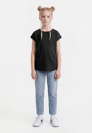 Ung pige stående, iført sort t-shirt, lyseblå jeans, hvide Nike-sneakers, med to flettede knolde og to frontfletninger.