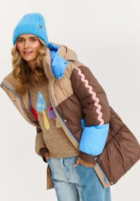 Žena v modrej pletenej čiapke, béžovo-hnedej prešívanej bunde s modrými manžetami, svetri s vzorom húb a svetlomodrých džínsoch, usmievajúca sa a pózujúca.