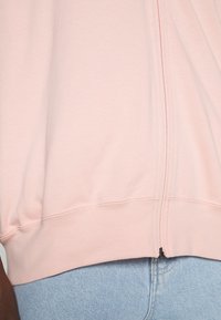 Detailní záběr na osobu nosící světle růžovou mikinu na zip a světle modré džínové kalhoty, ukazující zip a texturu látky.