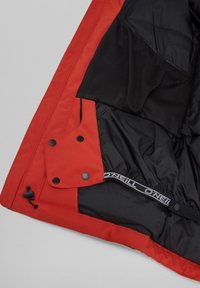 Veste à coque rouge avec doublure isolante noire, dotée de boutons-pression, de poches en mesh et d'une bande élastique avec logo au niveau de l'ourlet.