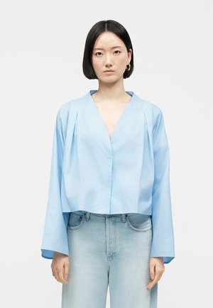 Giovane donna con capelli neri lisci che indossa una blusa azzurra chiara a collo a V e jeans a vita alta color blu sbiadito su sfondo bianco.