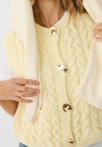 Żółta dziergana kamizelka z wzorem warkocza, z trzema dużymi złotymi guzikami z przodu. Na ramieniu zwisa jasnokremowy sweter.