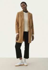 Brown wool-blend coat with notched lapels, single button closure, and flap pockets; worn over a cream knit sweater and black corduroy pants.