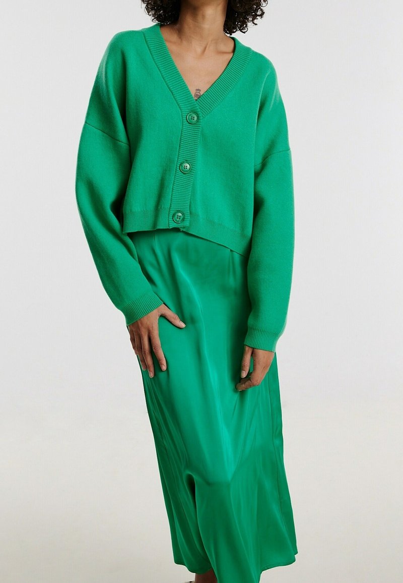Cardigan vert court avec un décolleté en V et une fermeture à boutons, associé à une jupe longue en satin vert lisse. Les deux vêtements présentent une texture douce.