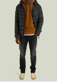 Sort pufferjakke med hætte, quiltet design, frontlomme med lynlås. Brun hættetrøje lagdelt under, parret med mørke jeans og beige sneakers.