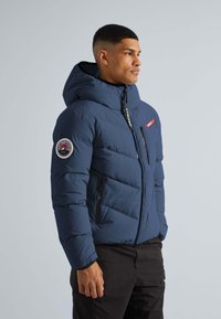 Marineblå pufferjakke med hætte, der har en lynlåslukning foran, to sidelommer og et cirkulært mærke på venstre ærme. Teksturiseret quiltet design.