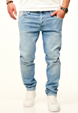 Man wearing light blue jeans, white sneakers, a white t-shirt, a wristwatch on the left wrist, and a bracelet on the right wrist.