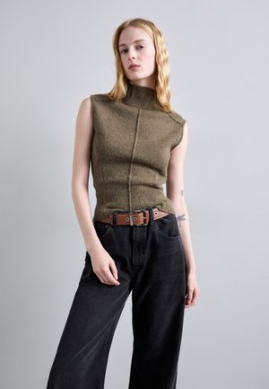 Jonge vrouw met lang blond haar, draagt een olijfgroene mouwloze coltrui, zwarte wijde broek en een bruine riem, staande tegen een grijze muur.