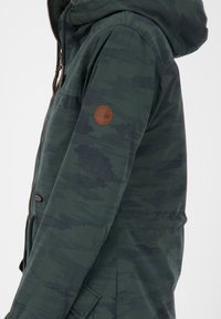Giacca verde a fantasia con cappuccio, caratterizzata da un logo rotondo marrone sulla manica sinistra, tessuto testurizzato e tasche frontali con zip.