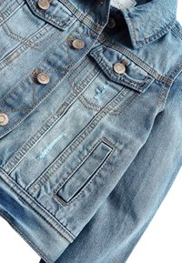 Veste en denim bleu clair avec des boutons en bronze, présentant des détails déchirés, deux poches poitrine et des coutures contrastantes.