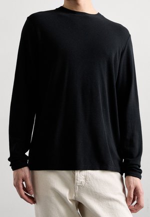 Person trägt ein schlichtes schwarzes Langarmshirt und hellbeige Hose, steht vor einem neutralen Hintergrund.