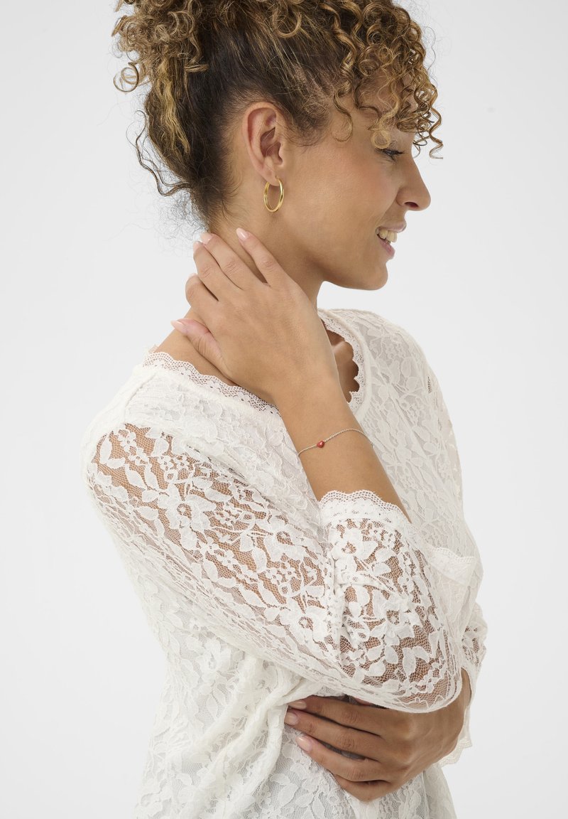 Spitzenoberteil mit langen Ärmeln, filigranen Blumenmustern, gewellten Kanten, ergänzt durch ein silbernes Armband mit einer roten Perle.