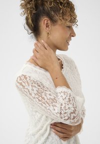 Blonde top med lange ærmer, intrikate blomster mønstre, bølgede kanter, suppleret med et sølv armbånd med en rød perle.